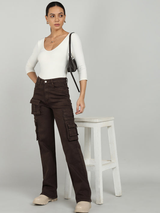 A woman wearing brown cargo straight-leg jeans from OMG Jeans, styled with a white fitted top, beige platform boots, gold accessories, and a black shoulder bag, posing next to a white stool.