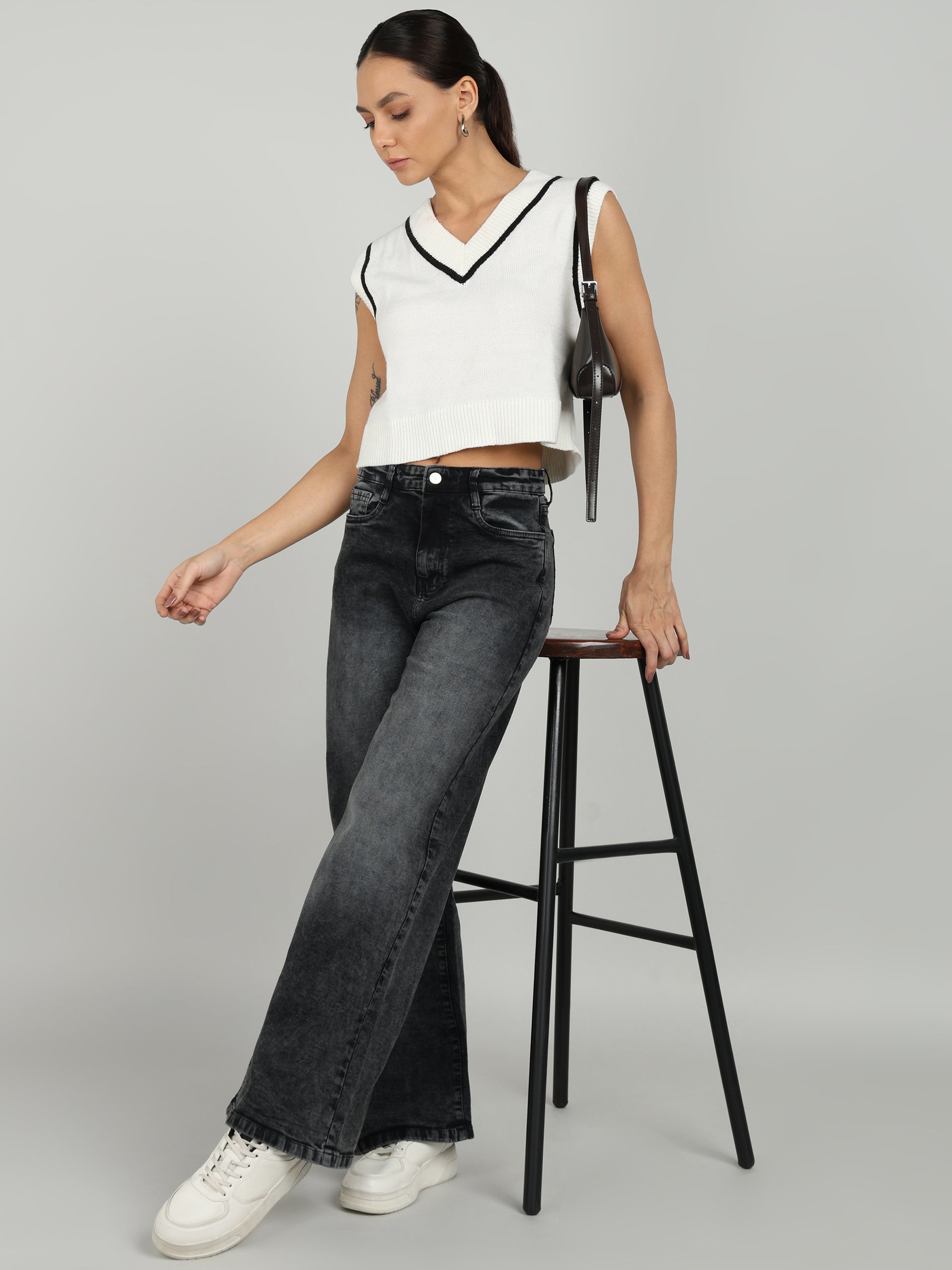 A stylish woman wearing black cargo pants from OMG Jeans, paired with a white sleeveless cropped sweater, white sneakers, and a black shoulder bag, posing confidently.