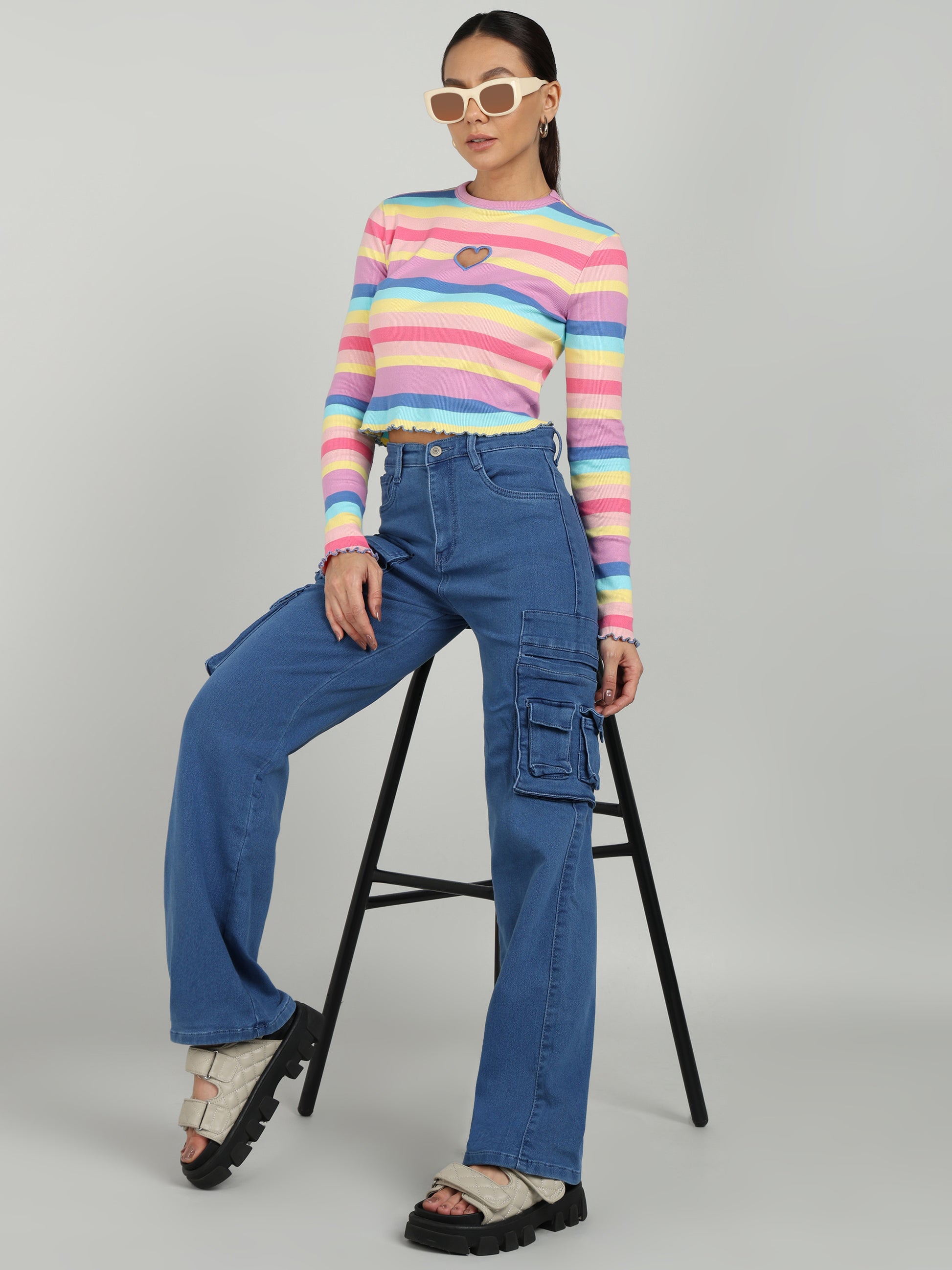 A woman wearing blue cargo wide-leg jeans from OMG Jeans, paired with a colorful striped crop top, chunky platform sandals, and white sunglasses, posing with a black stool.