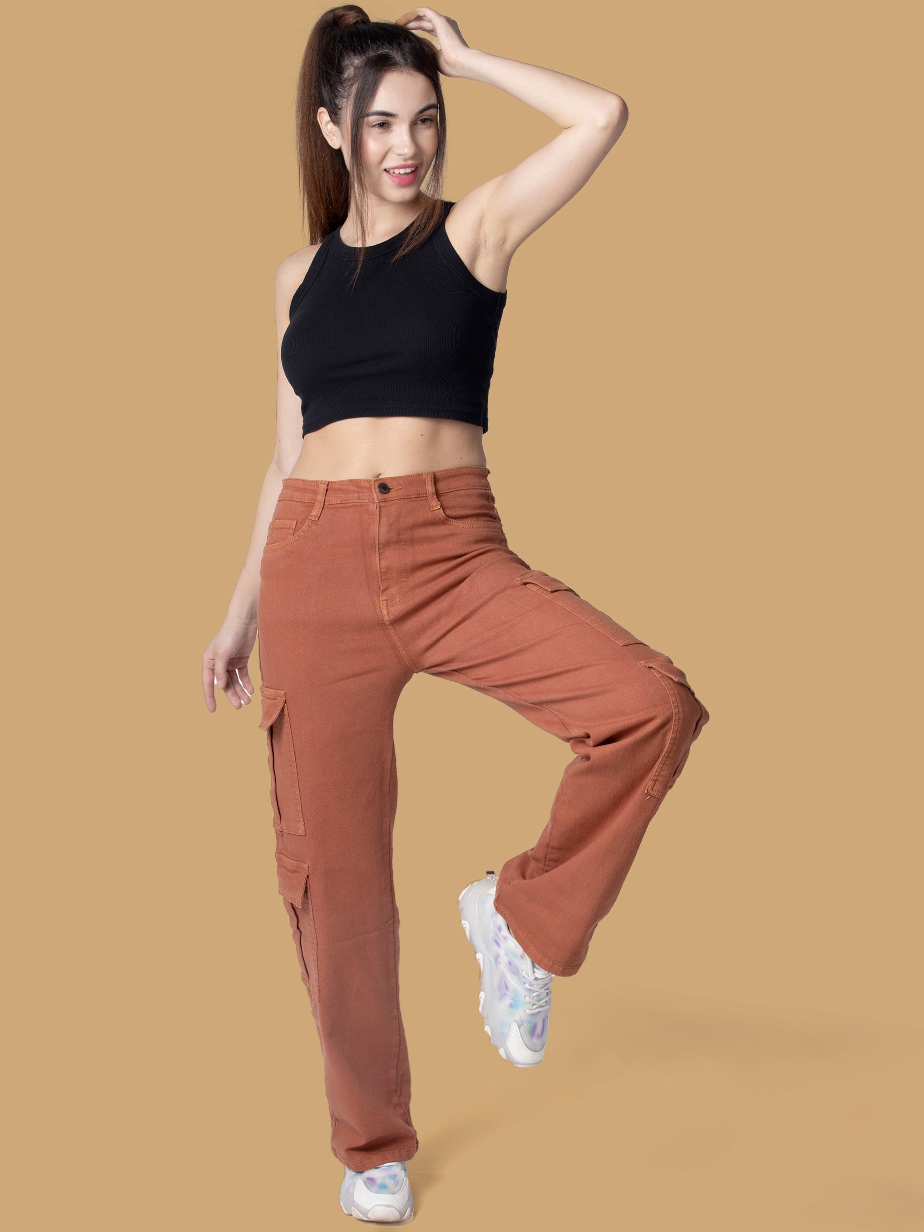 A trendy woman wearing brown cargo pants from OMG Jeans, styled with a black sleeveless crop top and white chunky sneakers, posing playfully against a beige background